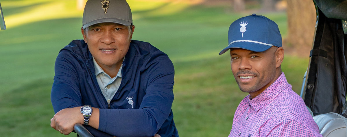 Two men in golf attire sitting in a golf cart on a grassy golf course. One man is wearing a navy blue polo shirt and shorts, and the other is wearing a pink checkered polo shirt, gray shorts, and white Nike golf shoes.